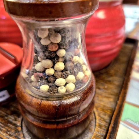 Photo of a partially see-through container with peppercorns of various color inside on a wooden tray and with other out-of-focus containers in the background.
