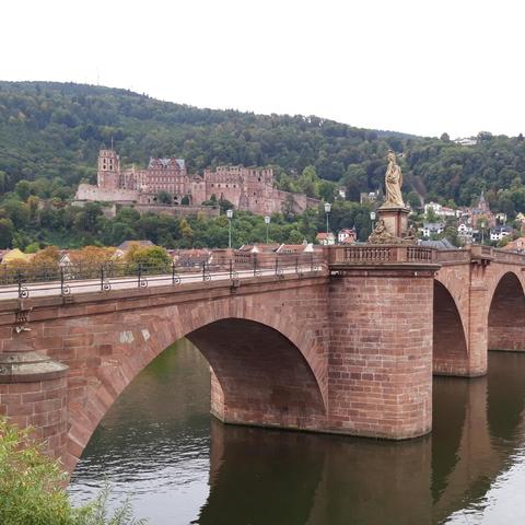 A red masonry arch footbridge spans a calm river, with a statue standing on one of its piers. In the background, a castle sits on a forested hillside above a town.