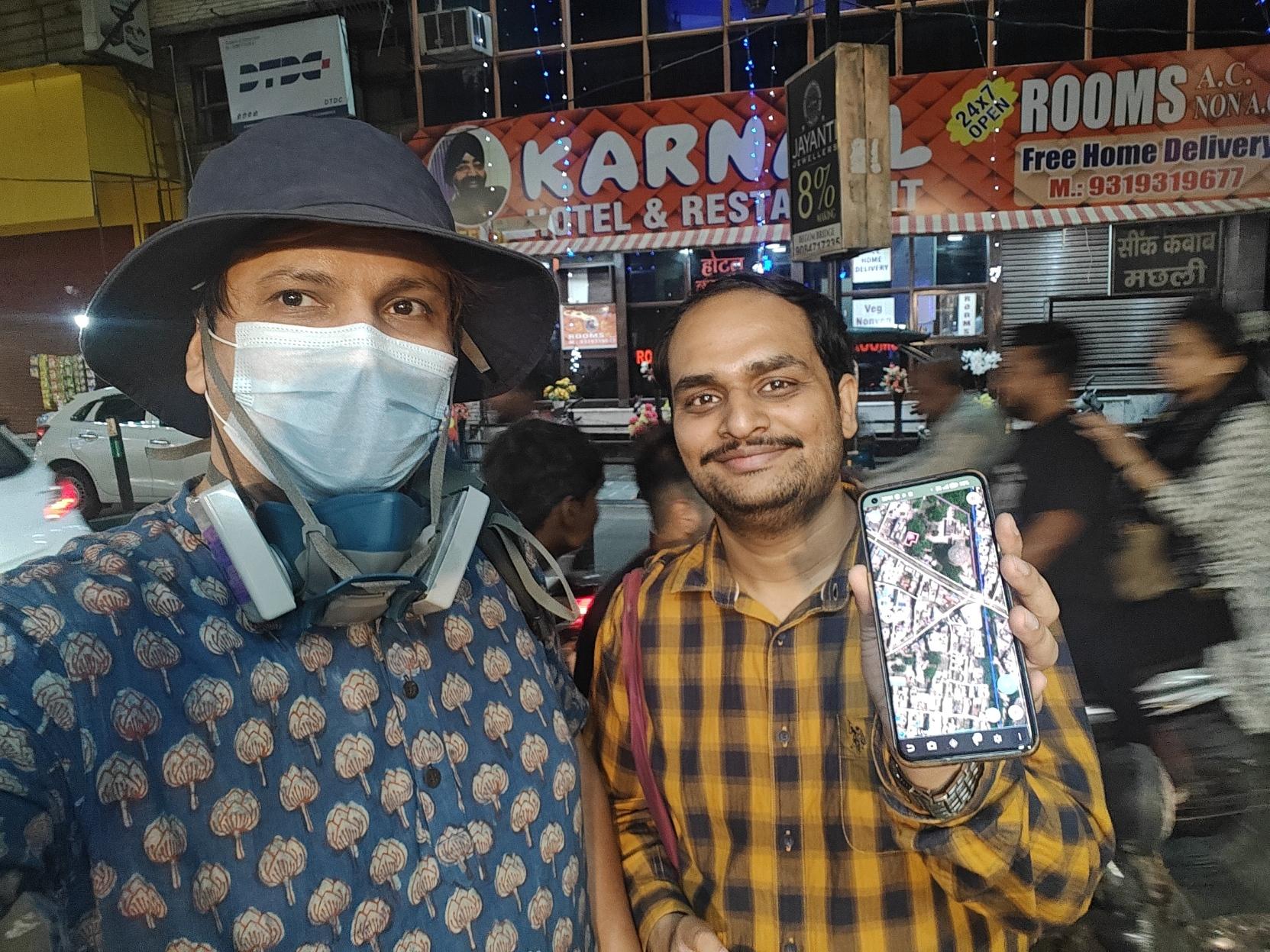 Selfie of two people standing on a busy streetside in Meerut. 

The one on the left is wearing a blue hat, a surgical mask, and a blue kurta with an Indian floral print. There's a blue elastomeric respirator hanging below per face.

The one on the right has a receding hairline, a moustache, and a slight beard, and is wearing a checked yellow full-sleeved shirt. Per is holding up a phone which has Vespucci open.