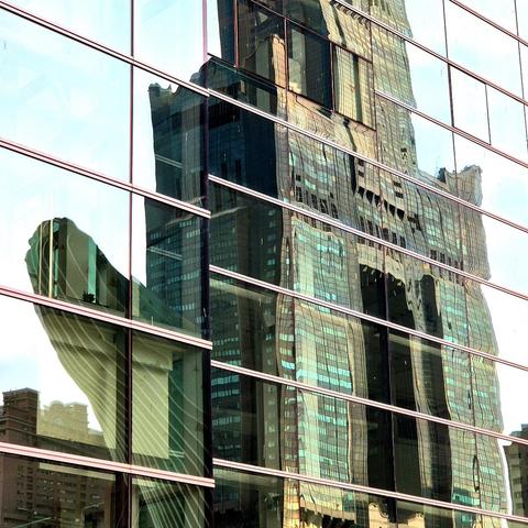 Glass window reflection of a tall skyscraper with green-tinted windows against a light-blue sky. The windows are broken up by horizontal metal frames.
