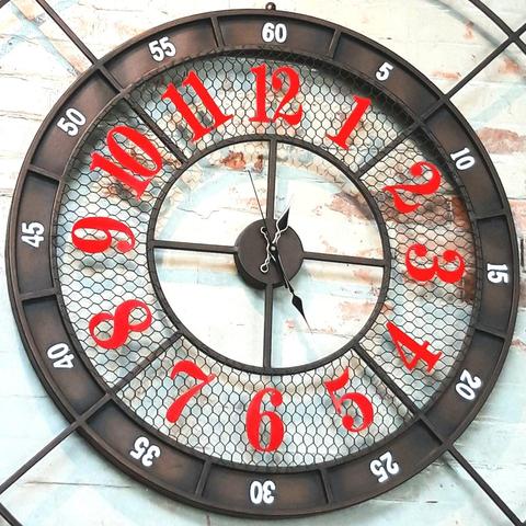 Industrial-looking metal clock featuring a narrow outer metal black band with 5-minute labels (5 to 60) in white sans-serif Arabic numerals, a wide middle mesh band with hour labels in bright red serif Arabic numerals, and an inner circular frame with cross hairs where the hour, minute, and second hands are mounted