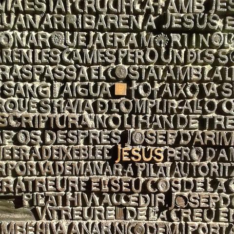 Rows of bronze raised uppercase letters forming words in various languages with the word “Jesus” highlighted in gold