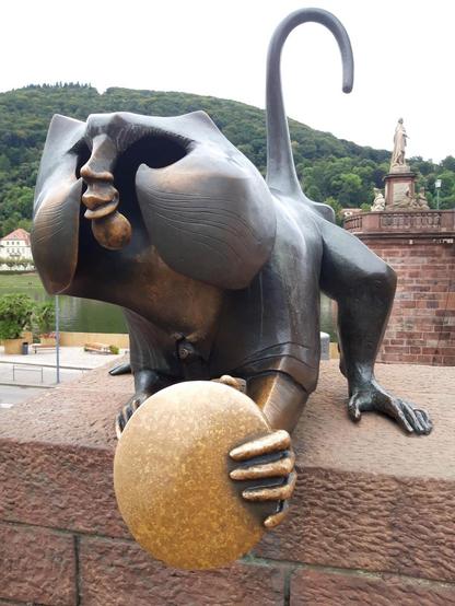 Bronze sculpture of a stylized monkey with a hollow head and holding a golden disk placed on top of a brick railing. Barely seen in the background are forested hills and a river and a portion of a bridge.
