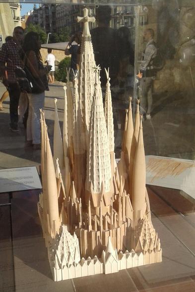 Scale model of the complete Basílica de la Sagrada Família in Barcelona looking towards its apse and inside a glass enclosure with reflections of tourists visible. The basilica is depicted with several tall curved pointed spires with a large cross on top of the tallest tower, a twelve-pointed star on top of the second-tallest tower, and winged subjects on the next tallest four.