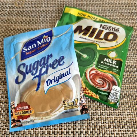 A light-blue plastic packet of instant coffee branded “San Mig Super Coffee” below which is “Sugar Free” in large letters followed by “Original” is smaller letters over a photo of a cup of coffee. Behind and partially covered is a green plastic packet of malt chocolate branded “Nestlé MILO” over a depiction of a cup of milk chocolate. Both packets are on top of a tan-colored woven-textured surface.