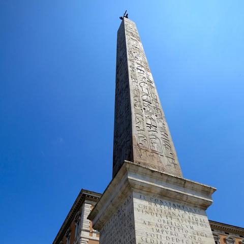 Tilted-up view of an Egyptian obelisk against a clear bright blue sky that gradually brightens to a light blue at the upper right corner. The side of the obelisk facing the sun is bright while the adjacent side is dark in shadow. The faces of the obelisk is covered in hieroglyphics. A top corner of a building is partially seen behind the obelisk.