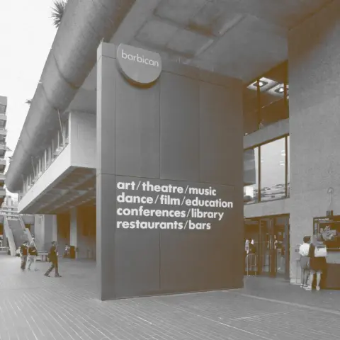 Monochrome photo with hints of sepia of a portion of brutalist building in front of which is a large freestanding two-story sign. The top of the sign has 3/4 of a circle at the top featuring the text, “barbican”, and in the middle is the following text:

art / theatre / music
dance / film / education
conferences / library
restaurants / bars