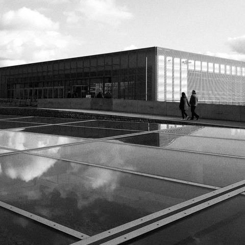 High-contrast black and white photograph of a stout rectangular building looking towards one of its corners. The façade facing left is in dark shadow while the side facing right is bright. In the foreground is a large array of horizontal glass panels installed near the ground and reflecting the building and the sky. Between the building and the panels is a footpath with two people holding hands walking towards the building.
