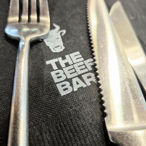 Photograph of a stainless steel cutlery set consisting of a fork, a steak knife, and a butter knife on a black paper napkin with the logo (cow head) and name of the restaurant (The Beef Bar) printed on it
