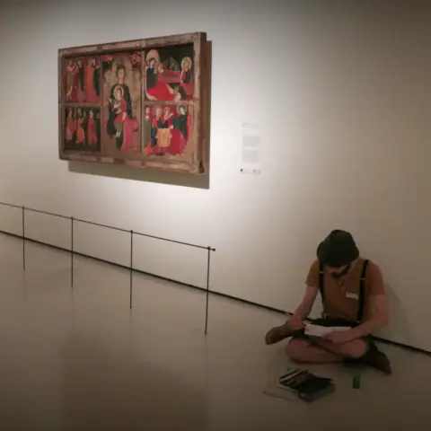 Museum scene featuring the Romanesque painting Altar frontal from Avià on a white wall with a very thin metal guardrail in front of it mounted on the floor. Sitting cross-legged on the floor against the wall near the painting is a man wearing a tan shirt, very dark shorts with suspenders, and a dark bucket hat with a set of color pencils on the floor in front of him. He is looking down on a sketch pad on his lap and drawing on the pad with an orange pencil on his right hand.
