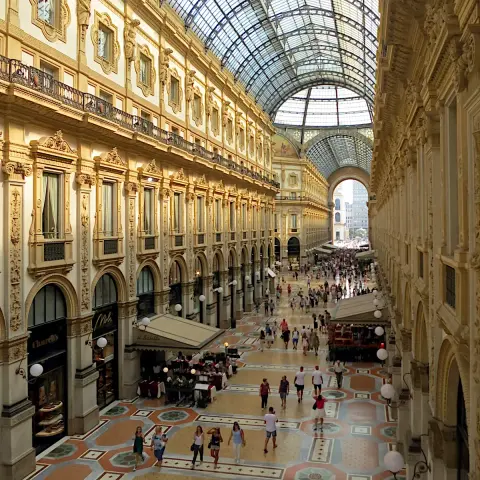 Interior view of a long multi-story Renaissance revival shopping arcade hall looking down from an upper floor. The arcade is covered with a curved glass ceiling and tourists and shoppers are on the ground floor. The scene is shot in the late afternoon with diffused sunlight pouring into the spacing giving the scene a warm glow.
