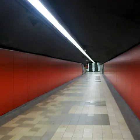 A moody photo of pedestrian tunnel with matte bold red paneled walls, square floor tiles in various shades of gray, and a very dark slightly curved ceiling with a single strip of continuous fluorescent lighting in the middle spanning the whole length of the corridor. In the distance, the corridor opens up to a wider space with a pair of escalators visible leading up to an upper floor. There are no people seen in the photo giving it a liminal space aesthetic.