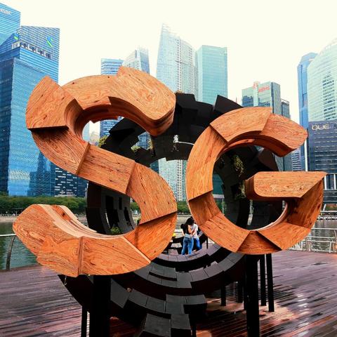 Wooden sculpture of the letters S and G with parts of each letter having different numbers of wooden layers. Both letters are mounted vertically on a horizontal pipe-like black-painted wooden mount with the same layered look as the letters and with the G slightly behind the S. The sculpture fills most of the 1:1 photo and is on a wooden esplanade with metal railings overlooking a body of water. On the far shore stands several modern skyscrapers behind which is a white overcast sky.