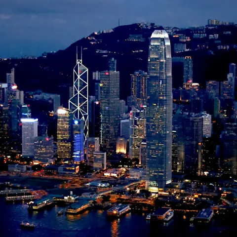 Blue hour photograph from a significant elevation of many skyscrapers between a body of water with several piers in the foreground and a dark hill range in the background at roughly the same height as the skyscrapers. The skyscrapers have many of their interior lights on and two tall skyscrapers are quite prominent: one is outlined in a very large triangular mesh of linear lights and topped with two antennas, while the other is the tallest in the view with a profile that gently tapers of toward…