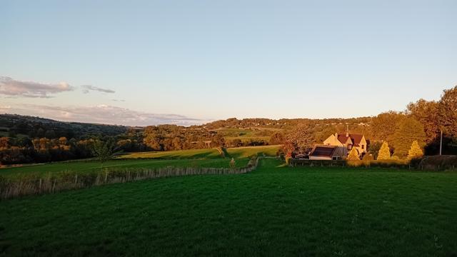 Rigardamte laŭ larĝa malprofunda valo for de malalta suno, kiu metas la proksiman kampon en ombro, sed lumigas la malproksiman arbaron per pala ora lumo. Ankaǔ estas farmdomo en la sunlumo ĉe la dekstra flanko de la foto. Ĉio sub pala blua ĉielo.