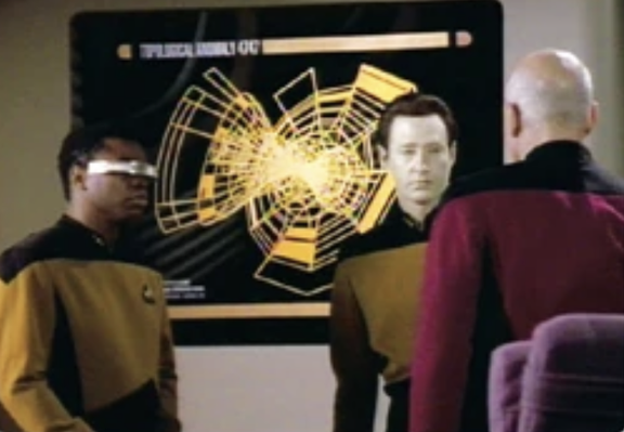 LaForge, Data, and Picard stand in front of a complex geometric shape shown on the wall screen in the main conference room of the Enterprise. Data is explaining things.
