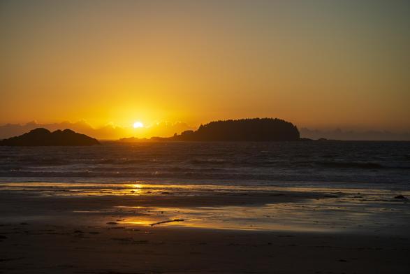 The sun sets behind a large cloud formation in the far distance, nearly behind the horizon. In the nearer distance, the silhouette of two islands is visible. The larger one has a few distinct buildings, as well as a lighthouse. The beach in the foreground is nearly flat, and the wet sands reflect the sunset light.