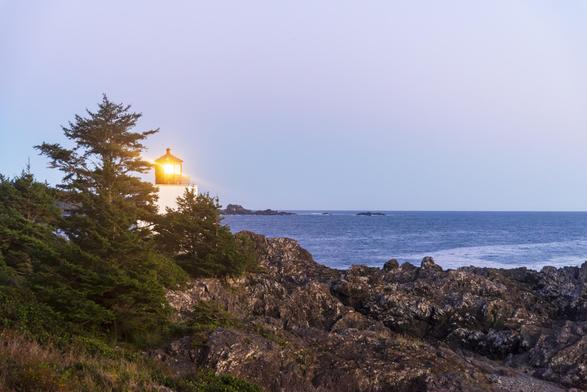 A lighthouse sits atop a large set of rocks in the shore, behind a few pine trees. It is right after sunset, and there is a purple cast in the landscape. The lighthouse beam shines exactly towards the camera, and so it glows with a bright orange color, and forms a glinting bright spot. The sea in the distance is wavy but otherwise featureless, just like the sky.