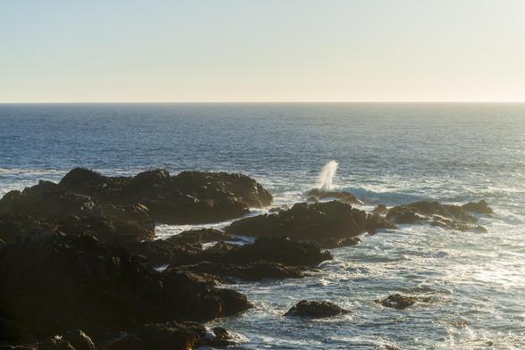 A large rocky formation is visible in the shoreline in the late day light. Waves crash against it, with spray shooting towards the air. The ocean surface is very wavy, but featureless otherwise, just like the sky.