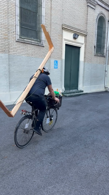 A guy on a fixed gear bike with a very narrow but long table strapped to his back like a backpack cycling out of frame