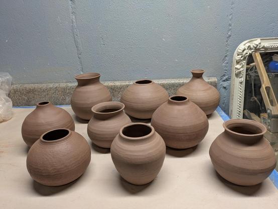 9 trimmed tan stoneware vases on a table in my studio. They have to dry completely before going in the bisque kiln.