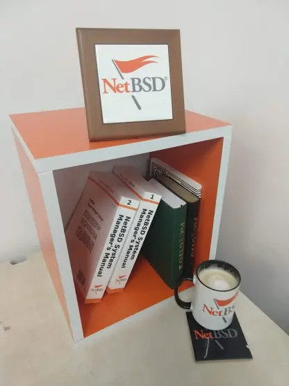 an orange and white cube shelf (only a single little cubicle) with a few books inside, including the NetBSD System Manager's Manual (printed manpages). also visible, a NetBSD mug with some coffee on a NetBSD ceramic coaster, and a little framed NetBSD logo on top of the shelf.