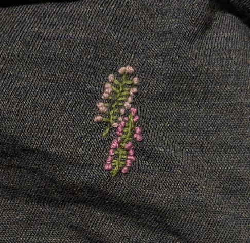 The two linear tears in the dark blue knit fabric have first been sewn together with green embroidery floss with blanket stitches on both sides of the tear, and then the ends of each of the "side branches" (i.e. the ends of the blanket stitches) have been decorated with purple and lavender-colored knot stitches to form the resemblance of small flowers.