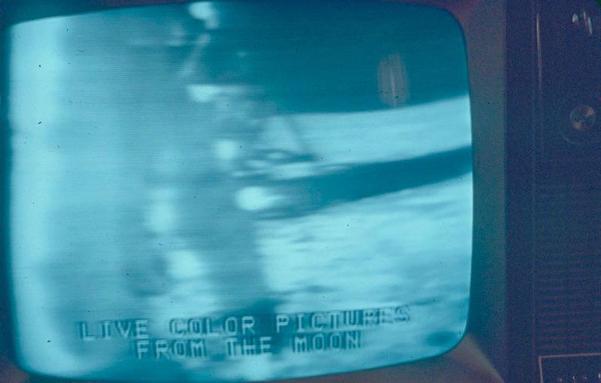 An old television screen displays a blurry image with the text "LIVE COLOR PICTURES FROM THE MOON," suggesting a historic broadcast of the moon landing. The image has a bluish hue, typical of early video transmissions.