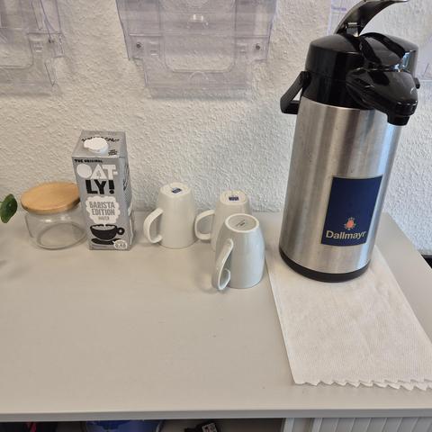 ein Sideboard mit einer großen Bürokanne Kaffee, drei umgedrehten, also noch nicht benutzten Tassen und einer Tüte Hafermilch.