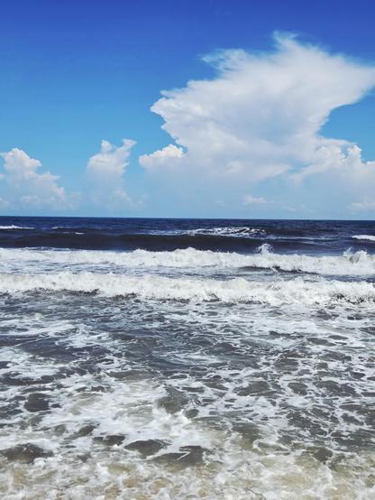 The Atlantic Ocean seen from Avon, NC, USA. The tide is in, minimal sand showing, and waves are lined up to crash on shore. The white foam hovers over gray water. Further out toward the horizon the water is a deep blue. The sky is a brightened blue littered with cumulonimbus clusters. An AI tool was used on this photo to erase a dude in the middle of the surf appearing to lose his trunks. It was too good an overall shot to have kept the photo bomber that wasn't immediately there when I triggered the shutter. I'm not sorry.