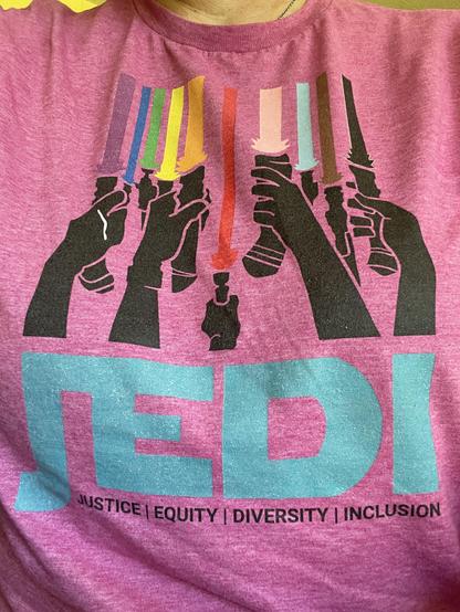 A pink t-shirt on a person with a logo of many black silhouette hands holding a rainbow of colored lightsabers. Underneath in Star Wars font is the acronym JEDI. Underneath, the words Justice, Equity, Diversity and Inclusion
