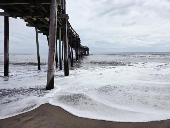 Snap of the Avon Pier, Avon, NC. The pier shot from the beach looking out under the pier toward the Atlantic Ocean. The pier starts out left of frame and gets smaller going out to sea in the center of the photo. The tide is coming in on the sand in large arcs of white foam. The sky is cloud covered making the ocean at the horizon look gray. Streaks of black sand are visible, possibly related to summer beach nourishment efforts in the area. The sand at the bottom of the photo looks like a solid surface with no shells or footprints visible as high tide has molded the beach with its force.
