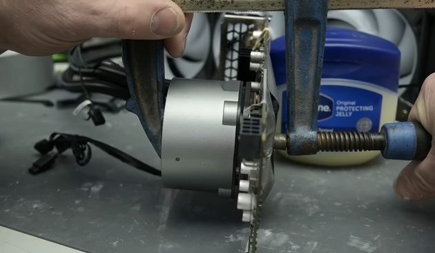 Picture showing a shop clamp being tightened to hold a water cooling heat sink onto a GPU card. The bottom side of the card is protected from the metal clamp by a 3D printed piece of plastic that is taped onto the card. This is actually the least interesting part of the video.