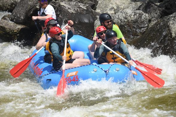 Five people wearing red helmets and a river guide in the back paddle a large blue inflatable river raft on a river churning with whitewater.