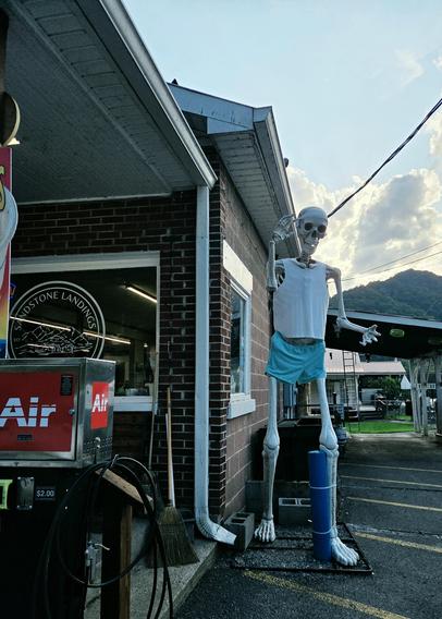 One of those 7 foot tall Home Depot skeleton yard decorations at the corner of a small gas station convenience store wearing blue shorts, a white tank top, and a sweat band around his skull. The skeleton is Right of frame with the rain spout on the store dividing the photo in the middle. Left of frame is the store front window reading "Sandstone Landings General Store" and the side of an air pump on the porch.