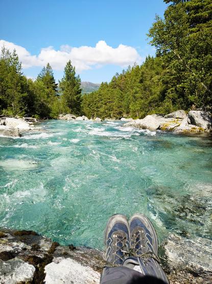 Ein Foto von einem schnellen Fluss, der durch eine felsige Landschaft fließt. Im Vordergrund sind die Füße einer Person zu sehen, die in Wanderstiefeln und dunklen Hosen steckt. Der Fluss ist türkisblau und weist an vielen Stellen weiße Gischt auf. Der Fluss fließt zwischen Felsen und um Bäume. Der Himmel ist blau und mit wenigen weißen Wolken bedeckt.