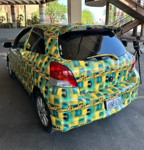 A photo from DefCon of a Toyota Yaris, viewed from the left rear, that has been covered with a pattern created by Bill Swearingen that defeats Flock cameras.  The pattern contains yellow and blue-green colors in a fuzzy square-ish pattern.