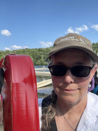 A person wearing a hot and sunglasses holding a red floating lifeguard tube with a tree lined lake behind and blue sky above e.