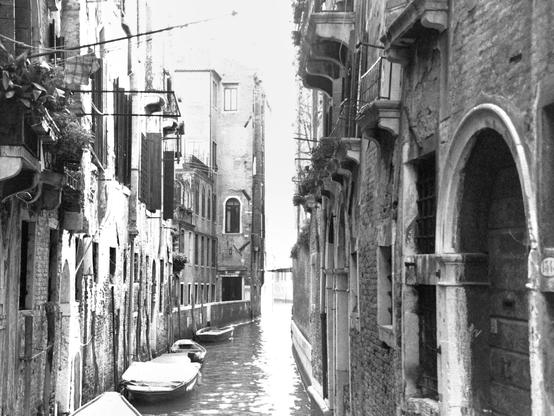 The black and white photograph, taken in a horizontal 4:3 format on film, shows a small canal in Venice, from a slightly elevated viewpoint, moored by several small boats,  Balconies, shuttered windows, arched doorways, and other details are visible on the old buildings on either side.