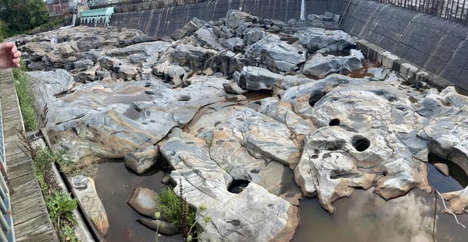 A panoramic view of the exposed riverbed below the dam on the Deerfield River at Shelborne Falls. It’s a weird landscape of irregular but smoothed rock with striations of light and dark grey, orange and white. Throughout, are circular holes varying in size from a foot to ten feet across and several feet deep. Many are filled with water and in some, the harder rock that ground out the hole is still visible in the bottom. A long angled concrete dam with a five foot wooden extension on top (that does not inspire confidence) extends across the top of the image. At far left are light green release gates and the spill way even farther along.