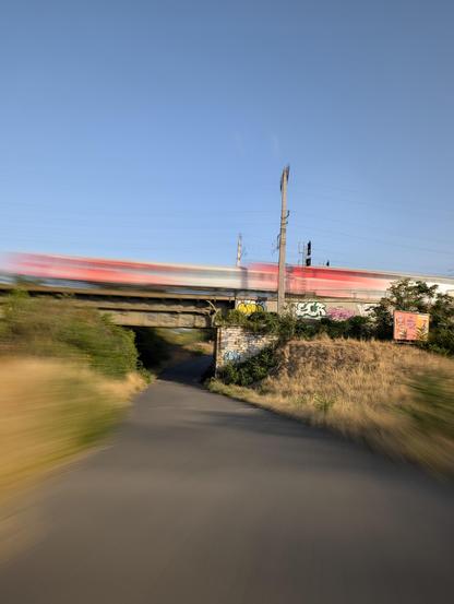 Foto einer Brücke, unter der eine Straße durch geht, oben auf der Brücke fährt soeben eine rot-weiße Eisenbahn von rechts nach links durch. Durch lange Belichtungszeit sind alle Bewegungen unscharf, der Zug von rechts nach links blurry und die Straße, auf der ich gerade fahre, sieht aus, als würde sie nach hinten zu uns gezogen werden.