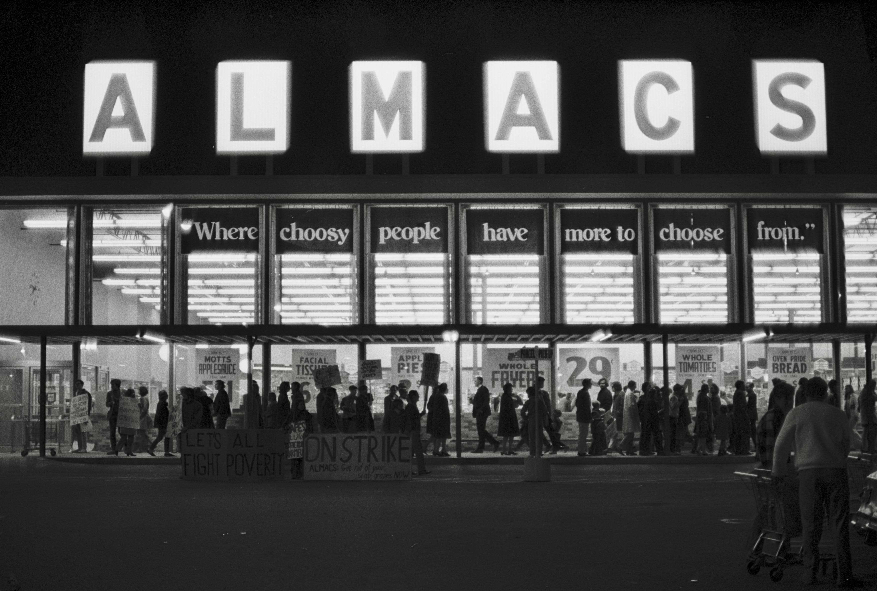Black and white night photo of a line of a picket line of people carrying signs in front of a brightly lit Almacs supermarket in Providence RI, 3 October 1969. The picket signs and banners say Boycott Almacs, Don鈥檛 Buy Scab Grapes, and other slogans, in support of an American Farm Workers Union harvest strike. The top of the supermarket window shows a backlit Almacs slogan: Where choosy people have more to choose from.