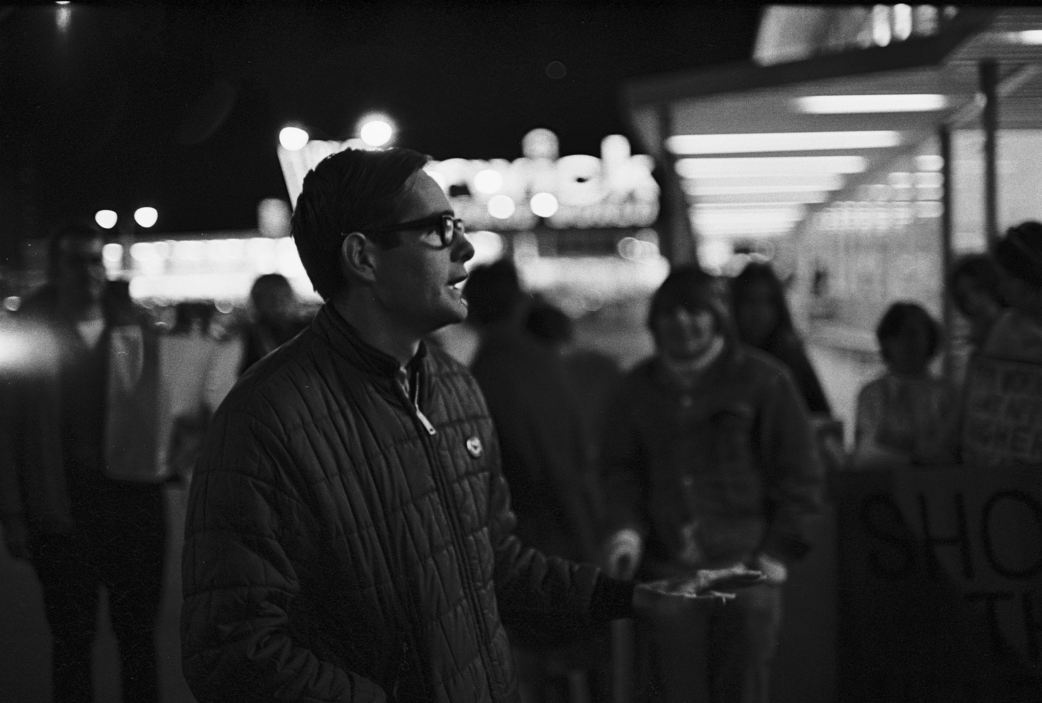 Black and white night photo of a protest leader speaking to grape boycott picketers lined up in front of an Almacs supermarket, Providence RI, 3 October 1969.