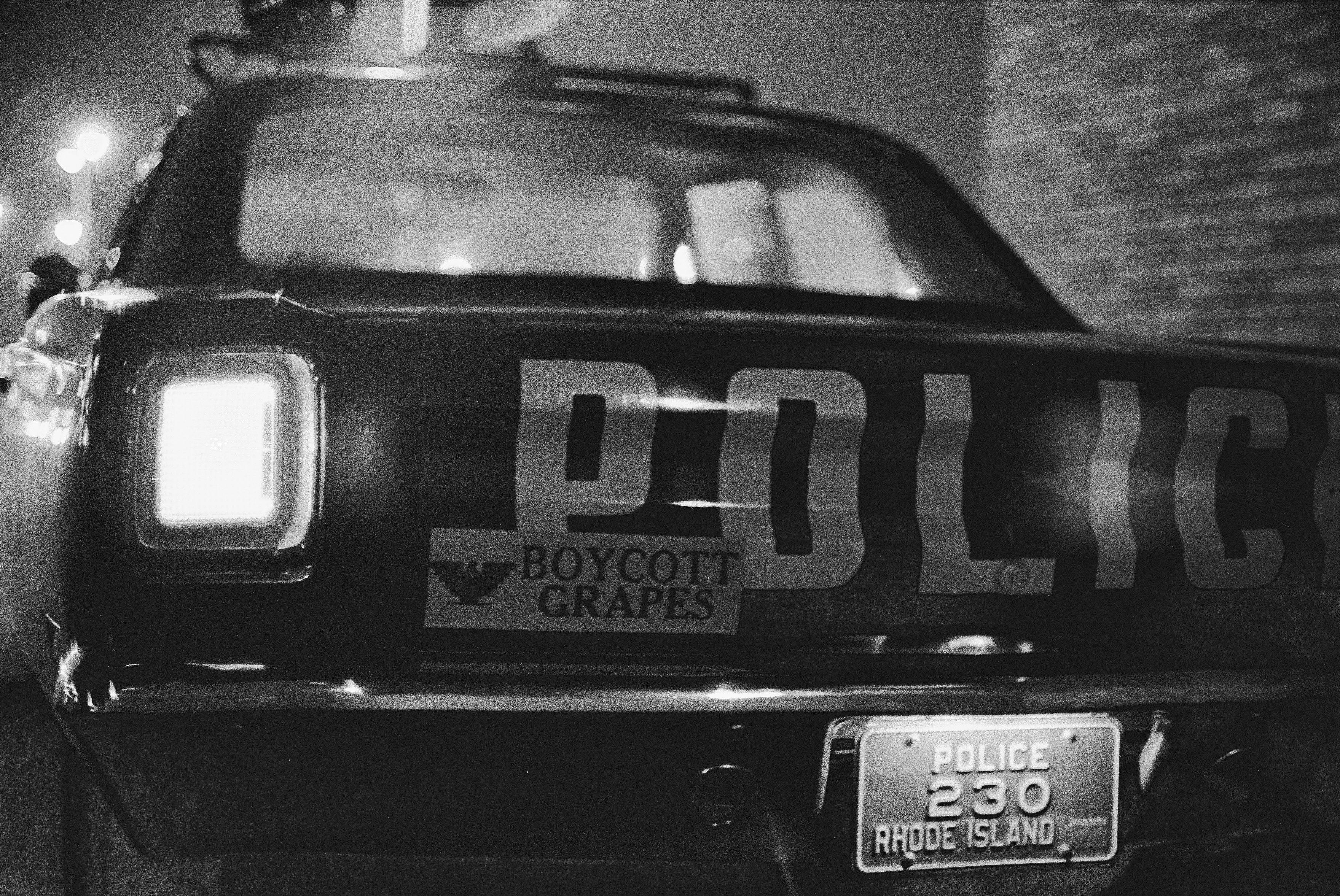 Black and white night photo of an empty Providence city police car, parked and idling with its lights on, in of an Almacs supermarket, Providence RI, 3 October 1969. The car has a Boycott Grapes bumper sticker with the American Farm Workers eagle covering part of the large POLICE painted on its trunk lid.