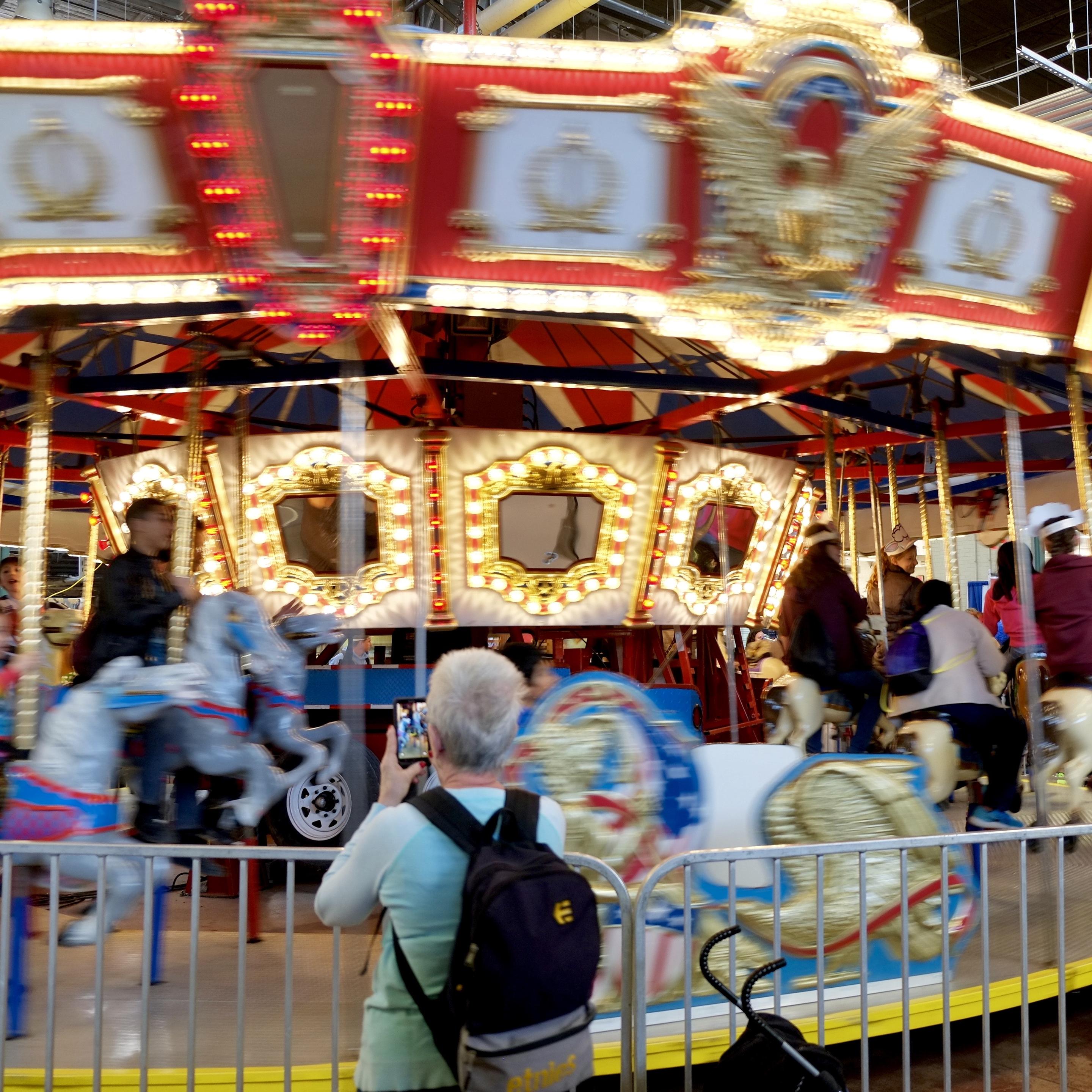 Color photo of a silver-haired woman taking a cell phone photo of a person riding a wooden horse on a carousel, as the person whirls into view from her left. An image of a person is clearly visible on woman’s cell phone screen. The top of the carousel is red with a large gold eagle and gold trim; below is a band of gold framed mirrors on a white background, each mirror surrounded by bright light bulbs. The people, horses, and a red, white, and blue sleigh on the carousel are blurred by their motion as the carousel spins to the right.
