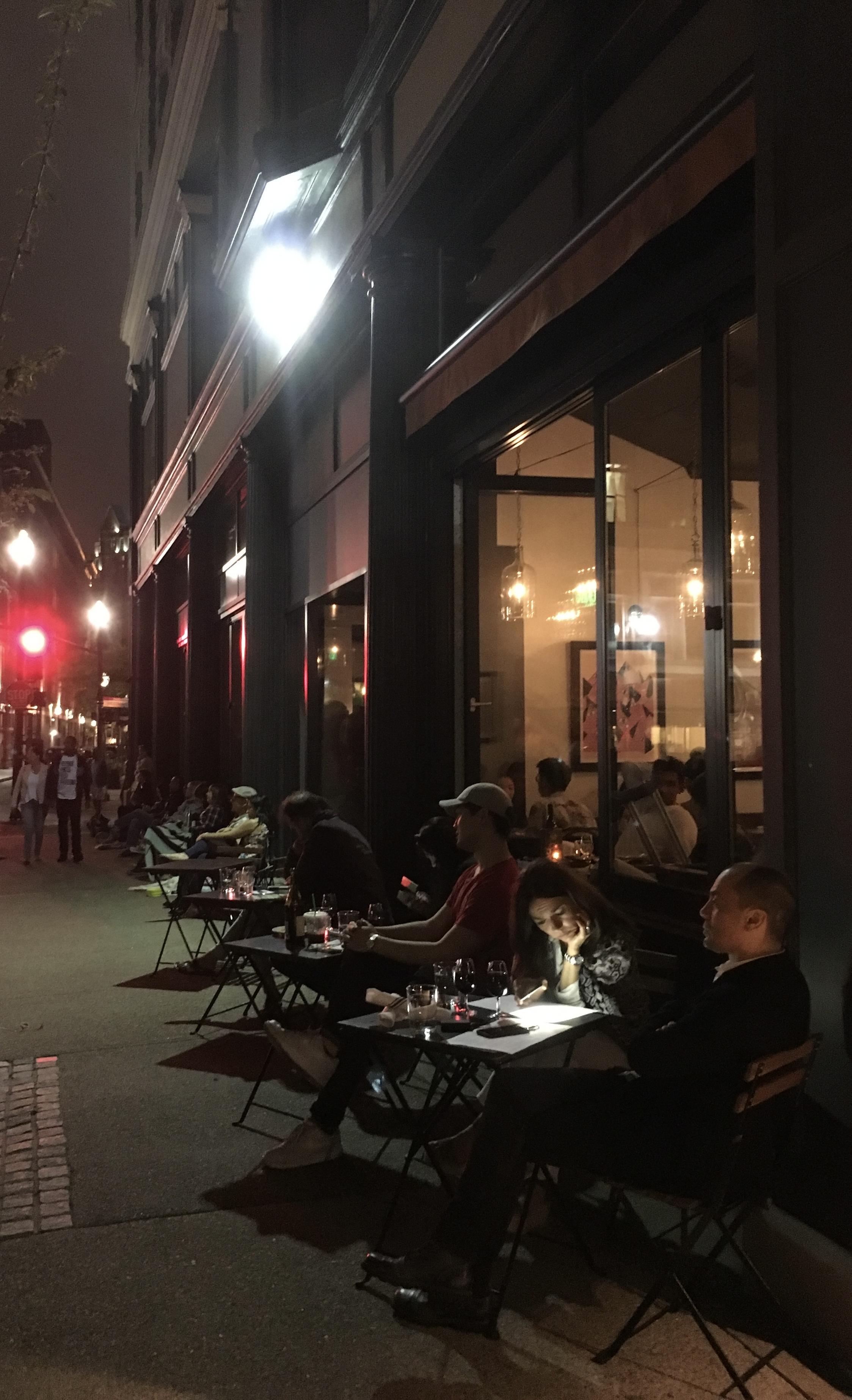 Color night photo of people eating, drinking, and talking at small cafe tables outside the windows of the Oberlin restaurant at its original location. At 9pm in June, the sky is dark, but with a some reflected city glow. A red traffic light and bright 19th street lamps on the sidewalk ahead light a couple walking towards the restaurant. In the foreground, a couple sit at a table set with wine glasses and printed single page menus. One of them leans forward, using her cellphone to illuminate her menu, which reflects light back onto her pensive face.