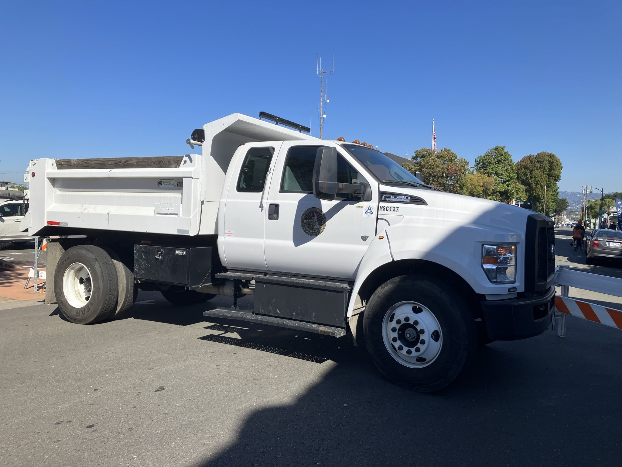 City street scene with a white dump truck parked perpendicular to the traffic lanes. The truck has a city seal on the passenger door. There are some trees, a parked car, an antenna tower, and a US flag on a flagpole in the background.