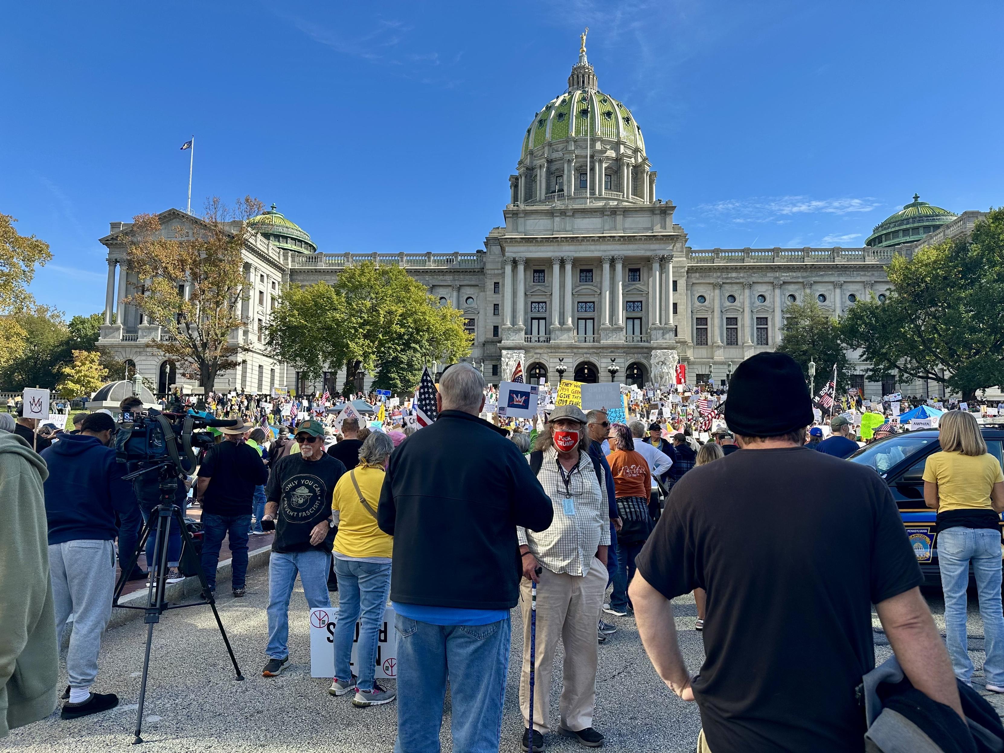 A TV news crew and small crowd of No Kings protesters apparently waiting to be interviewed stand across the street from a large colorfully dressed crowd of No Kings protesters who stand on the steps of the Pennsylvania State Capitol building, Harrisburg PA. The protesters carry a wide variety of homemade and printed No Kings signs. It’s noon on a sunny October day. A glittering gold statue tops the green Capitol dome under a bright blue sky