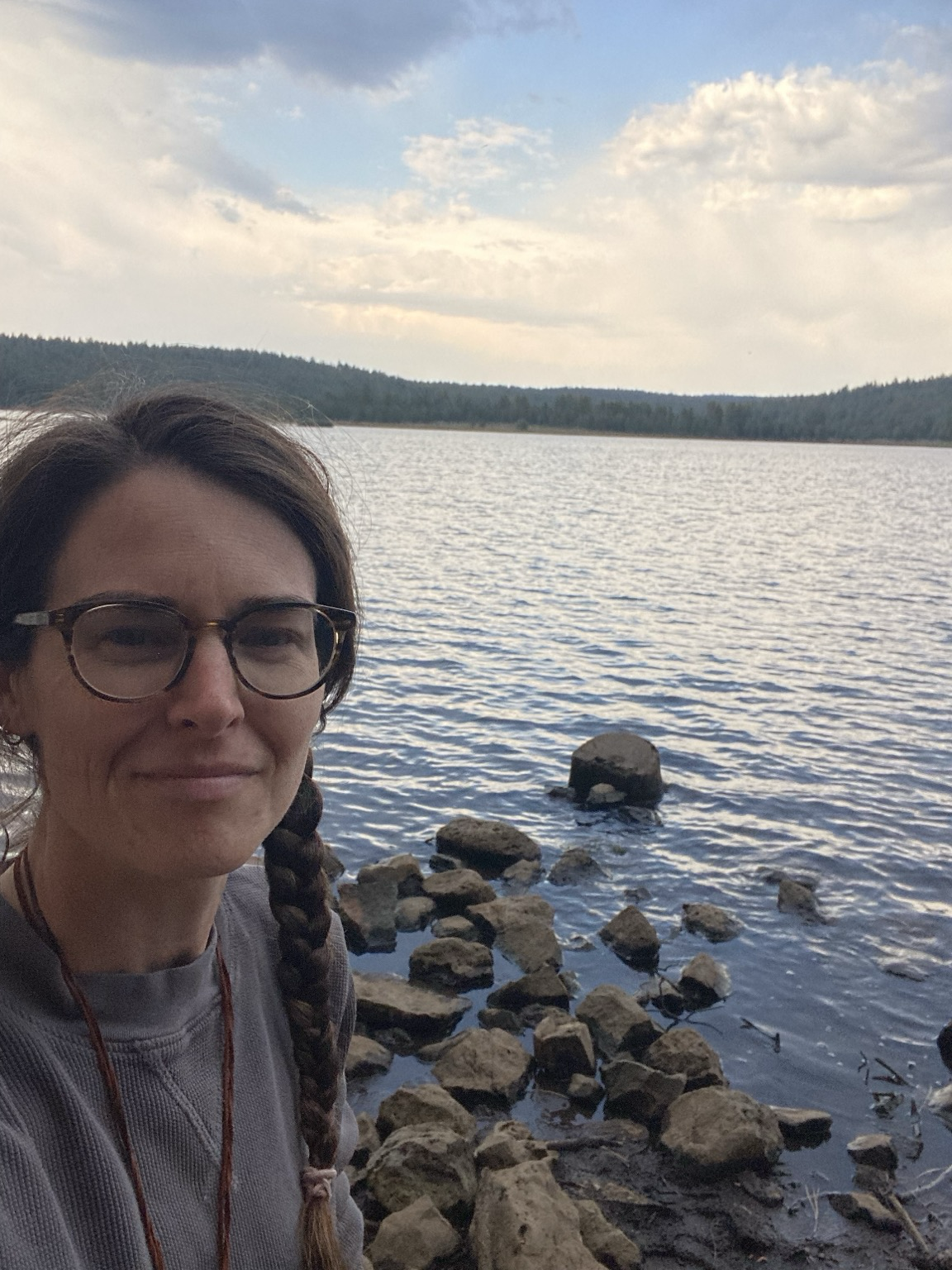 Selfie of a Person with glasses and a braid in front of a lake with trees dotting the far shore