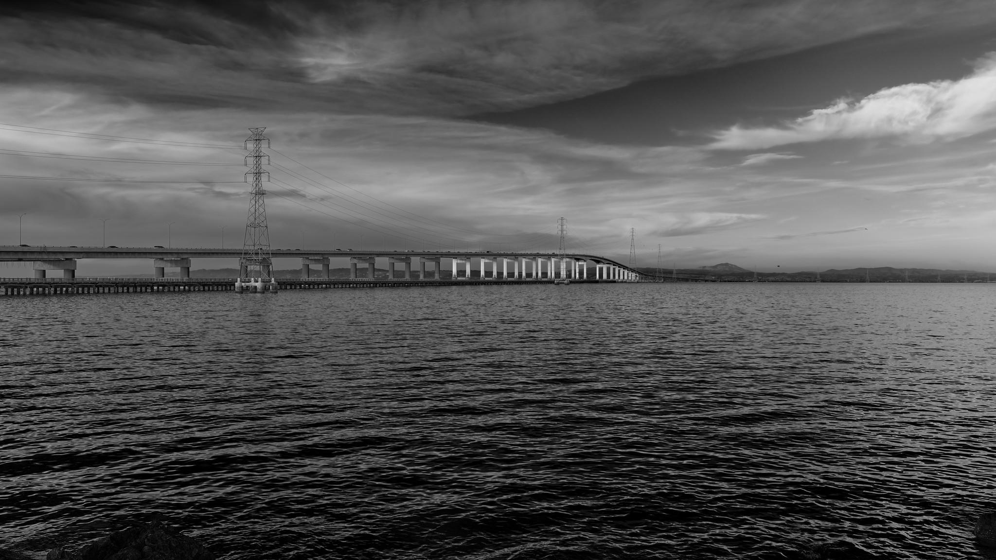 A very long but otherwise relatively unremarkable bridge crossing a large body of water. Pylons for high voltage power lines run alongside the bridge.